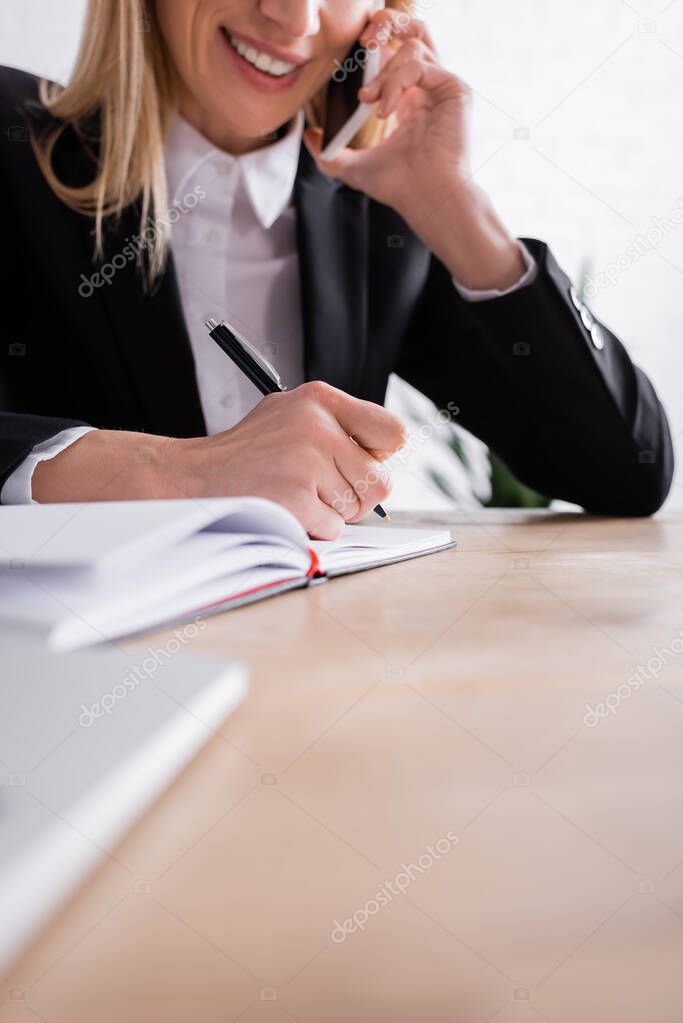 partial view of smiling notary talking on smartphone and writing in notebook on blurred foreground
