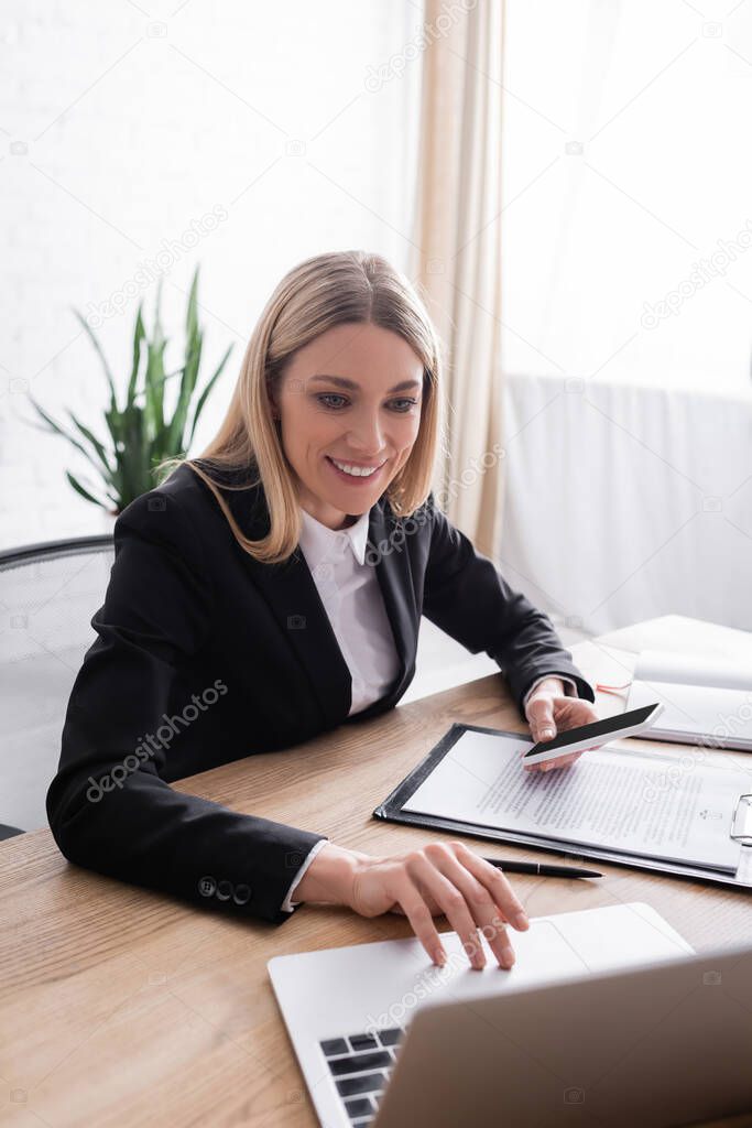 cheerful lawyer using laptop while holding smartphone near clipboard 