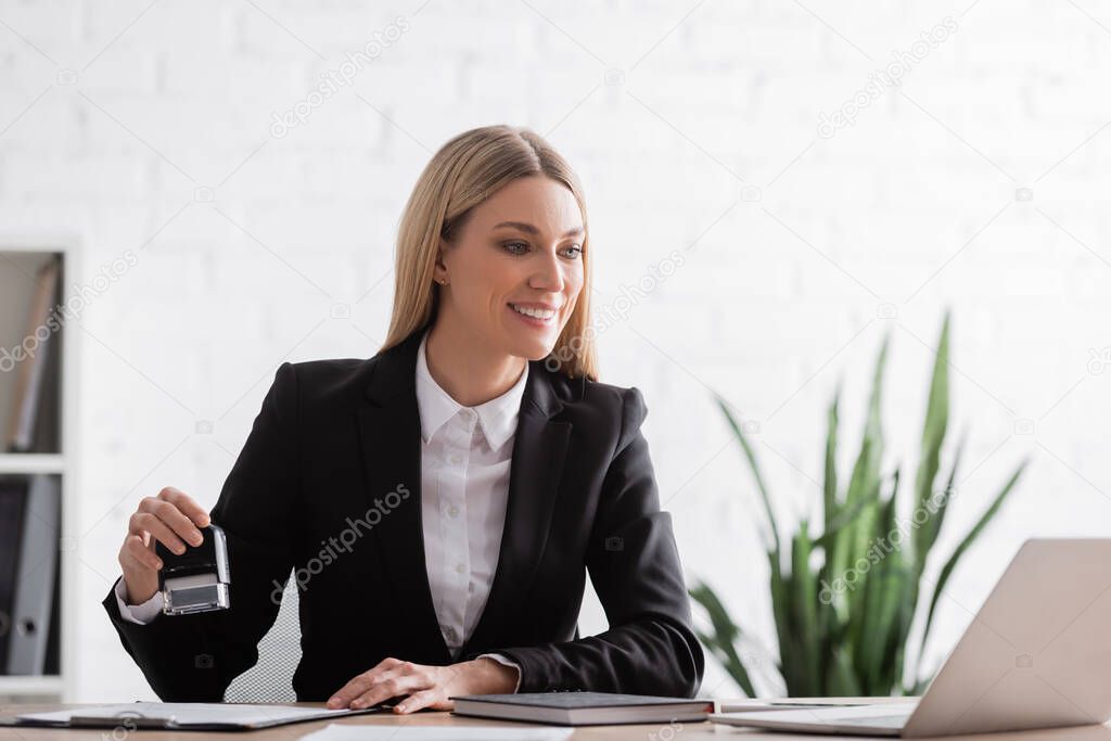 smiling notary in formal wear holding stamper and looking at laptop in office