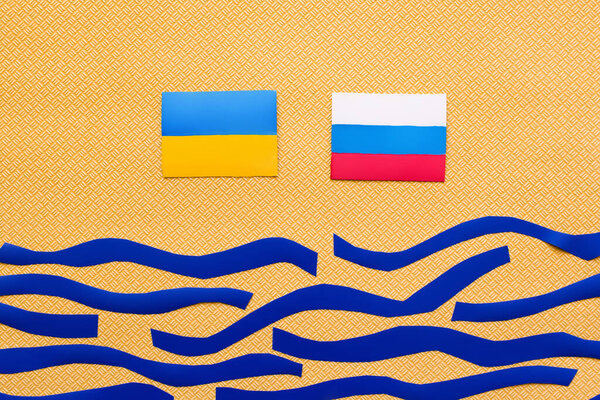 Top view of Ukrainian and russian flags above paper sea on textured yellow background 