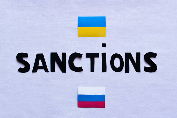 Top view of sanctions lettering near ukrainian and russian flags on white background 