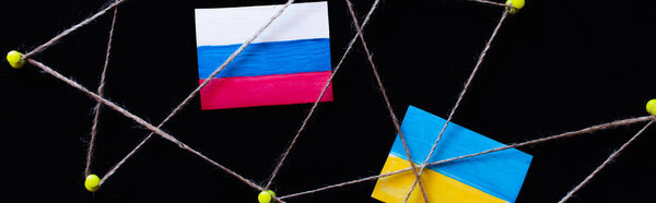 Top view of ukrainian and russian flags near push pins with thread isolated on black, banner