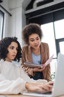 African american businesswoman holding paper folder near arabian colleague using laptop 