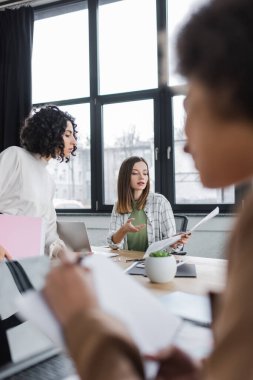 Businesswoman pointing at newspaper near laptop and muslim colleague in office 