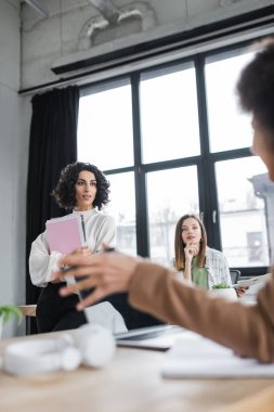 Interracial businesswomen with papers looking at blurred african american colleague in office 