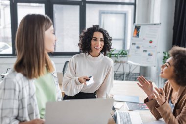Positive muslim businesswoman holding cellphone near interracial colleagues in office 