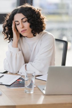 Tired arabian businesswoman sitting near clipboard and laptop in office 