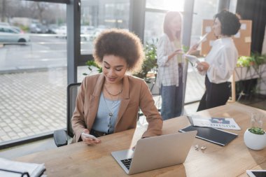 Smiling african american businesswoman using cellphone and laptop near newspaper and earphones in office 