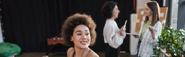 Cheerful african american businesswoman looking away near blurred colleagues in office, banner 