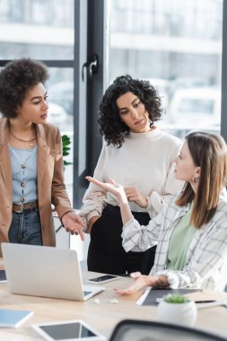 Blurred businesswoman pointing with hand near multiethnic colleagues and gadgets in office 