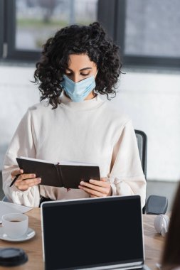Medikal maskeli Arap iş kadını dizüstü bilgisayarının yanında not defteri ve ofisinde kahve tutuyor. 