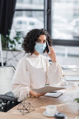 Tıbbi maskeli Müslüman iş kadını cep telefonuyla konuşuyor ve evrakları ofisinde tutuyor. 