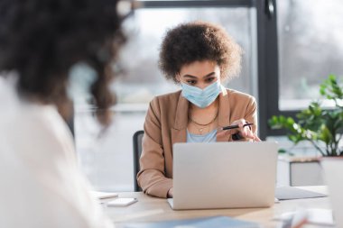 African american businesswoman in medical mask holding pen near devices and blurred colleague in office 