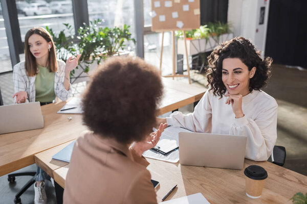 Cheerful muslim businesswoman looking at blurred african american colleague near devices and coffee in office 