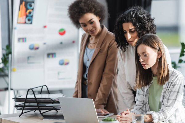 Young businesswoman looking at laptop near blurred multiethnic colleagues in office 