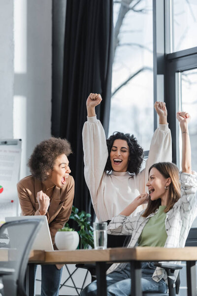 Excited multiethnic businesswomen showing yes gesture near laptop on table in office 