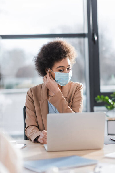African american businesswoman in medical mask using earphone and blurred laptop in office 