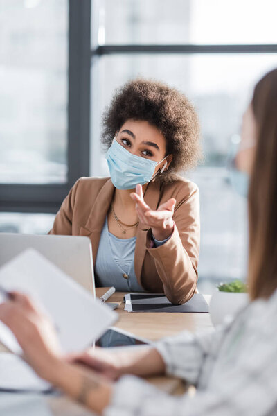 African american businesswoman in medical mask pointing at blurred colleague 