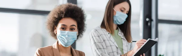 African American Businesswoman Medical Mask Earphones Looking Camera Blurred Colleague — стоковое фото