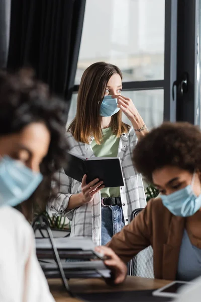 Young Businesswoman Holding Notebook Wearing Medical Mask Blurred Multiethnic Colleagues — Stockfoto