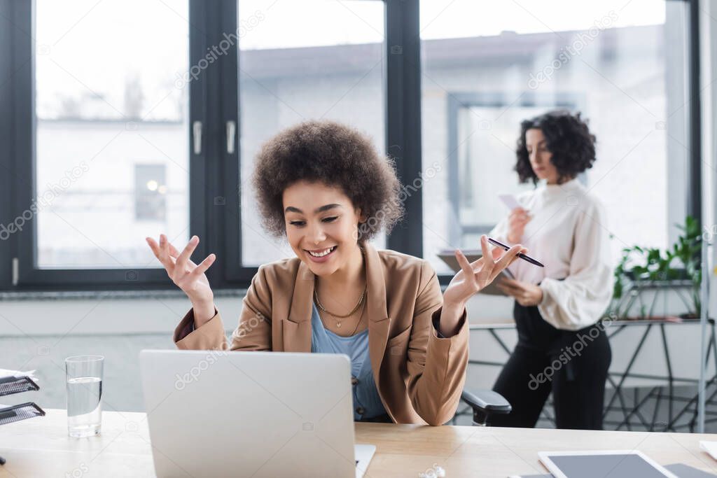 Cheerful african american businesswoman having video chat on laptop near blurred colleague in office 
