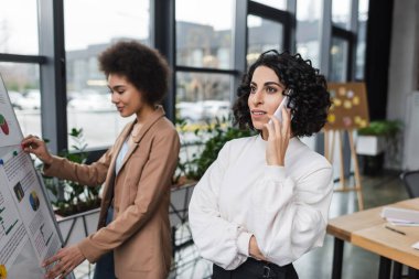 Genç Arap iş kadını, bulanık Afrikalı Amerikalı meslektaşının yanında akıllı telefondan konuşuyor ve ofis çizelgesini çeviriyor.