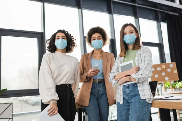 Międzyrasowe Biznesmenki Maskach Medycznych Trzymające Urządzenia Dokumenty Biurze — Zdjęcie stockowe