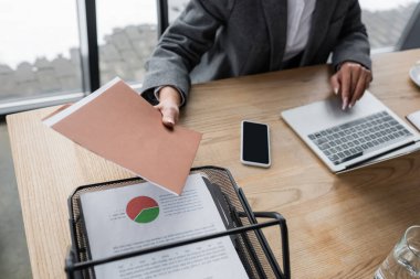 partial view of financier holding folder near smartphone with blank screen while working at laptop