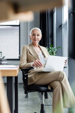 successful banker smiling at camera while sitting with laptop and smartphone on blurred foreground