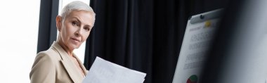 senior businesswoman with documents looking at camera near blurred flip chart, banner