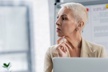 stylish senior banker looking away while thinking near blurred laptop