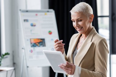 smiling banker looking at digital tablet near blurred flip chart with infographics