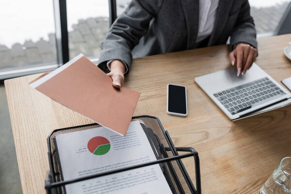 partial view of financier holding folder near smartphone with blank screen while working at laptop