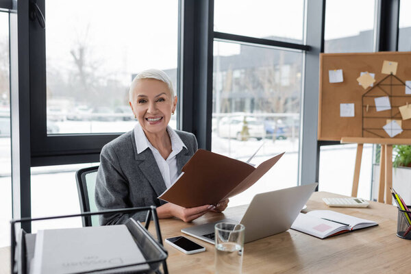 happy banker with folder smiling at camera near laptop and cellphone with blank screen