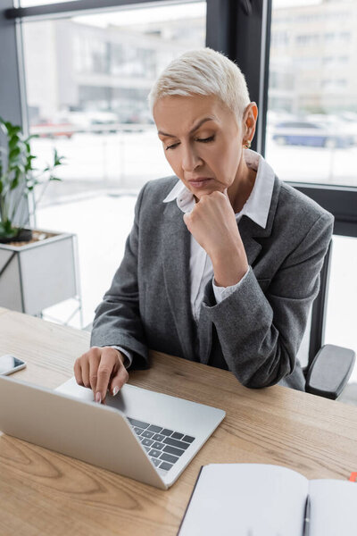 thoughtful banker holding hand near face while working on laptop in office