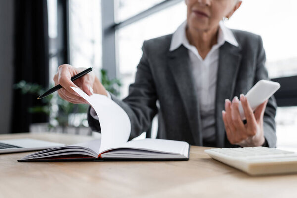 partial view of blurred banker with smartphone and notebook in office