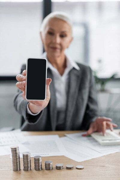 banker showing smartphone with blank screen near silver coins and documents on blurred background
