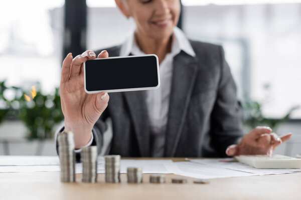 cropped view of smiling banker holding smartphone with blank screen near coins, blurred background