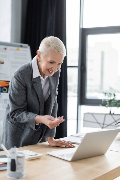 smiling banker gesturing near laptop during video call in office