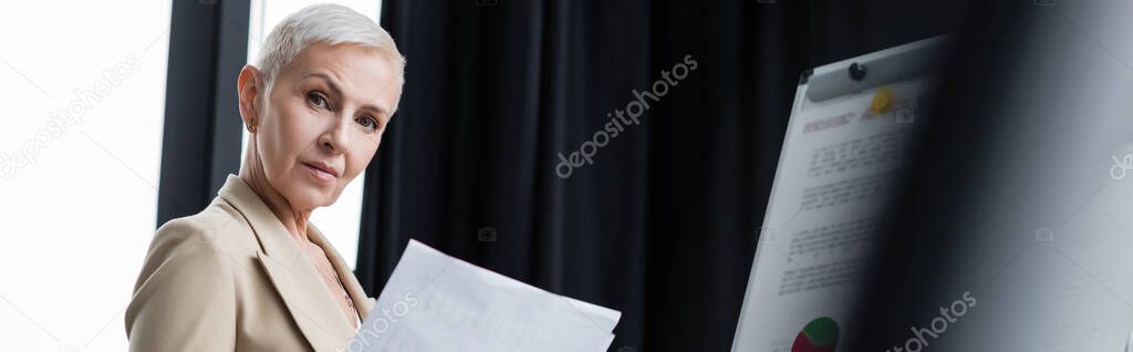 senior businesswoman with documents looking at camera near blurred flip chart, banner