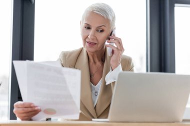grey haired banker talking on smartphone while holding papers near blurred laptop