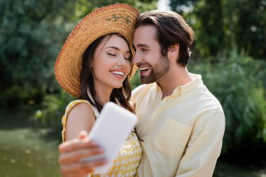 Hasır şapkalı mutlu genç kadın dışarıda neşeli erkek arkadaşıyla selfie çekiyor.