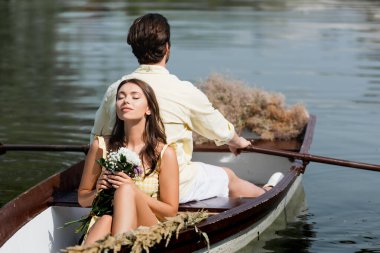 Genç bir kadın, elinde çiçeklerle romantik bir tekne gezisinde erkek arkadaşının sırtına yaslanıyor. 