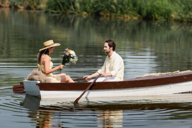 Hasır şapkalı, elinde çiçekler olan ve adamla romantik bir tekne gezisi yapan genç bir kadın. 