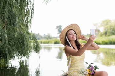 Hasır şapkalı ve elbiseli mutlu genç bir kadın gölde tekne gezisinde selfie çekiyor.