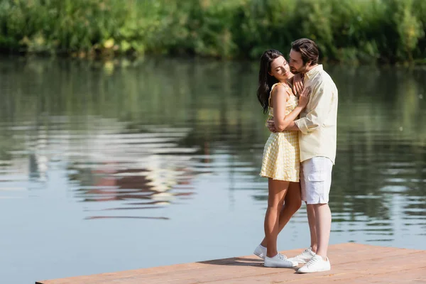 Yaz kıyafetleri içinde göl kenarındaki iskelede sarılan genç bir çift.