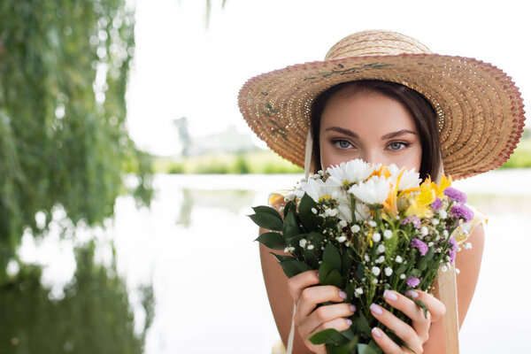 young woman in straw hat covering face while smelling flowers 