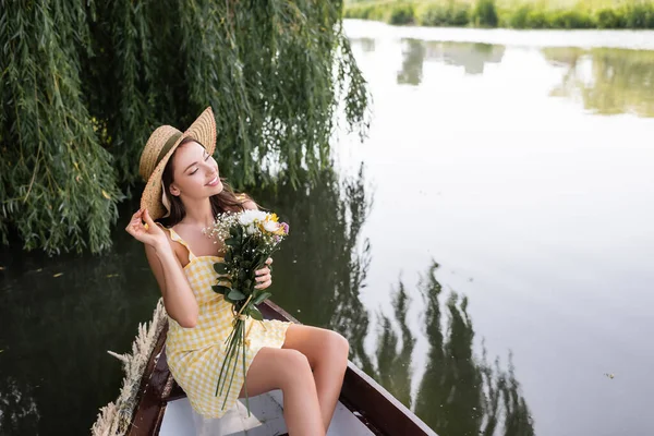 夢のような若い女性が藁帽子を調整し 湖でボートに乗っている間に花を保持 — ストック写真