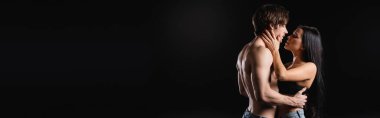 Side view of passionate brunette woman touching shirtless boyfriend isolated on black, banner 