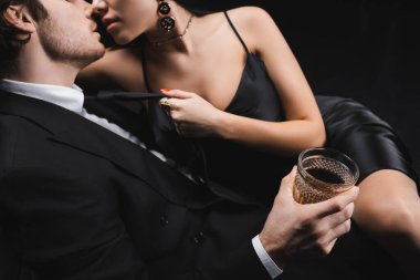 Cropped view of man in suit holding whiskey and kissing sexy girlfriend in silk dress isolated on black 